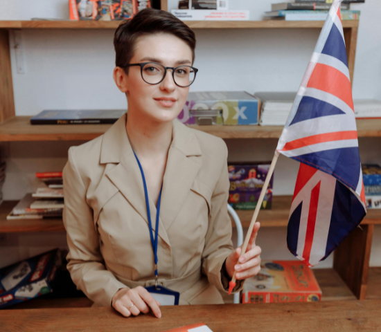 Young woman with glasses, wearing a beige blazer, holding a small British flag while sitting at a desk with books and games in the background.