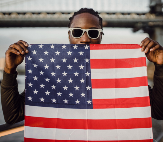 Man wearing sunglasses holding up an American flag in front of him, with a bridge in the background.