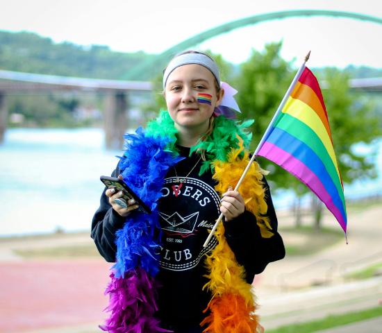 Person at Pride event wearing colorful feather boa, face paint, and holding rainbow flag, with bridge and water in background