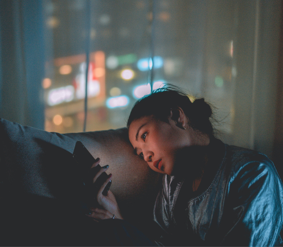 Person using smartphone at night, illuminated by screen light. City lights visible through window in background.