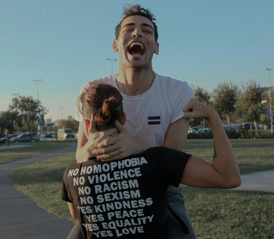 Two people embracing, one wearing a shirt with social justice messages including 'No homophobia, No violence, No racism'. Person in front laughing joyfully.